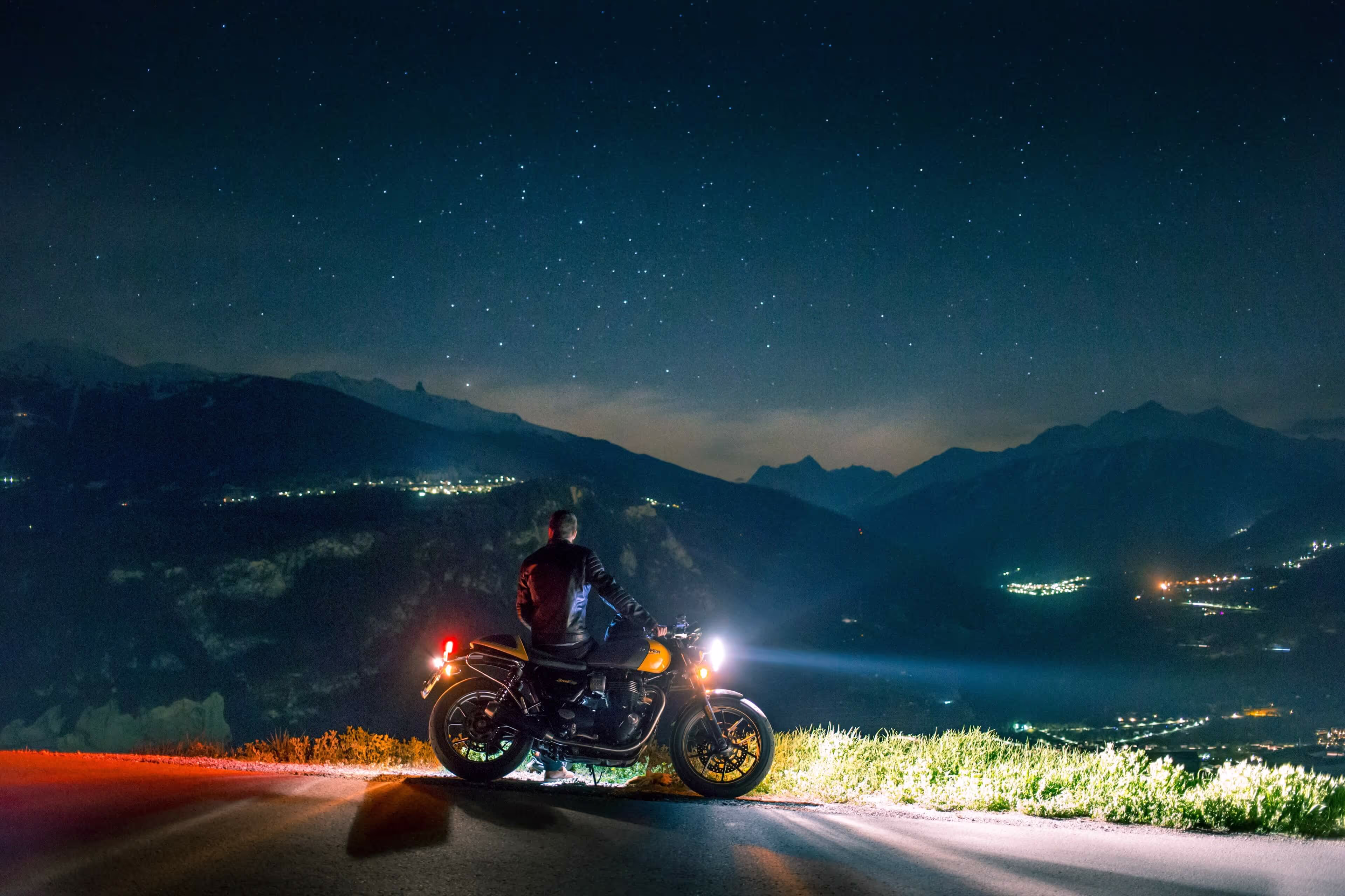 A man sits on a motorcycle on the side of a road, overlooking a valley and city at dusk.